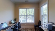 This single-floor space functions as a dedicated business center or office area, thoughtfully designed to support productivity in a comfortable environment. The room features workstations with granite or stone-topped desks positioned along the walls, each equipped with computer setups and ergonomic seating, making it well-suited for focused work or research tasks. A large, prominently placed window fitted with white horizontal blinds serves as the focal point of the space, flooding the interior with generous natural light and offering pleasant views of lush green trees beyond. An additional window on the adjacent wall further enhances the bright, airy atmosphere. The walls are finished in a warm neutral tone, complemented by classic crown molding that adds a touch of refined detail. Patterned carpeting in muted tones covers the floor, contributing to a professional yet inviting ambiance. A decorative plant adds a touch of greenery to the interior, reinforcing the connection to the natural surroundings visible through the windows. Overall, the space strikes a balance between functional utility and comfortable design, making it a welcoming environment for work or study.