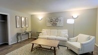 This single-floor space exudes a calm, welcoming ambiance through its carefully considered design and neutral tones. The living area serves as the centerpiece of the space, featuring soft taupe and warm beige walls that create a soothing backdrop for the furnishings. A plush white sofa and matching armchair are arranged around a rustic wooden coffee table, offering a comfortable and stylish seating area ideal for relaxing or entertaining. A patterned area rug in muted blue and cream tones anchors the space, adding subtle texture and visual interest to the hardwood-style flooring beneath. Warm wall sconces provide soft, ambient illumination throughout, complementing a sleek floor lamp that adds to the layered lighting scheme. The walls are adorned with carefully selected artwork, including a striking Buddha-inspired canvas that serves as a focal point, alongside framed feather prints that reinforce the space's tranquil aesthetic. A decorative console table along one wall offers additional display space, styled with a sunburst mirror accent. The textured ceiling adds architectural character, while the overall design language of the space balances simplicity with warmth, resulting in an environment that feels both polished and deeply relaxing.