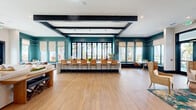 Located at Ascend Village in Winter Garden, Florida, this single-floor community space offers a generously sized, open-plan interior designed with a modern coastal aesthetic. The space is anchored by striking teal accent walls that contrast beautifully against crisp white ceilings accented with bold black exposed beams, creating a visually dynamic architectural statement throughout.

Light wood-tone flooring extends across the entire space, lending warmth and continuity between the distinct functional zones. A long communal bar-height counter with woven rattan seating serves as a central gathering and coworking area, bathed in natural light from a full wall of floor-to-ceiling windows that frame lush palm tree and waterfront views beyond. Roman shades provide adjustable light control throughout.

A cozy lounge area near the entrance features plush seating, a vibrant area rug, decorative leaf wall art, and a wall-mounted display screen. Built-in window seat benches offer additional casual seating alongside intimate dining tables. Pendant lighting with woven black fixtures adds a refined, resort-inspired touch. A private room with glass-panel doors is accessible off the main floor, providing an enclosed space for focused use. The overall ambiance is bright, welcoming, and thoughtfully designed for both social and productive activities.
