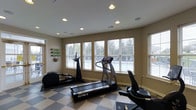 The Glenmary Village fitness center is a well-appointed, single-floor exercise space designed to support an active lifestyle. The room is bright and welcoming, benefiting from a row of large windows fitted with white blinds that allow ample natural light to filter in while offering views of the surrounding greenery outside. The ceiling features recessed lighting, ensuring the space remains well-lit at all times of day. Underfoot, a bold checkered tile floor adds a classic visual element and provides a durable, easy-to-clean surface suited for a fitness environment. The room is equipped with a selection of cardio machines, including a treadmill, an elliptical trainer, and a stationary bike, catering to a variety of workout preferences. A wall-mounted yoga mat rack adds to the room's versatility, signaling that the space accommodates both equipment-based and floor-based exercise routines. The neutral wall tones and clean layout contribute to a focused, distraction-free atmosphere. Overall, this fitness center offers residents of Glenmary Village a convenient and comfortable space to maintain their health and wellness routines.