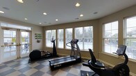 The Glenmary Village fitness center is a well-appointed, single-floor exercise space designed to support an active lifestyle. The room is bright and welcoming, benefiting from a row of large windows fitted with white blinds that allow ample natural light to filter in while offering views of the surrounding greenery outside. The ceiling features recessed lighting, ensuring the space remains well-lit at all times of day. Underfoot, a bold checkered tile floor adds a classic visual element and provides a durable, easy-to-clean surface suited for a fitness environment. The room is equipped with a selection of cardio machines, including a treadmill, an elliptical trainer, and a stationary bike, catering to a variety of workout preferences. A wall-mounted yoga mat rack adds to the room's versatility, signaling that the space accommodates both equipment-based and floor-based exercise routines. The neutral wall tones and clean layout contribute to a focused, distraction-free atmosphere. Overall, this fitness center offers residents of Glenmary Village a convenient and comfortable space to maintain their health and wellness routines.