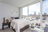 This contemporary studio unit offers a thoughtfully designed open-concept layout across a single floor, seamlessly blending living, sleeping, and dining areas into one cohesive space. Light wood-style flooring runs throughout, complementing the clean, neutral palette of soft gray and white tones on the walls and ceilings.

The living and sleeping area is the heart of the unit, anchored by a large floor-to-ceiling window that floods the space with abundant natural light and frames an impressive panoramic view of the city skyline. The open kitchen lines one wall and features a full suite of black appliances — including a refrigerator, electric range, and over-the-range microwave — paired with dark lower cabinetry, crisp white upper cabinets, and a light-toned countertop, creating a bold two-tone contrast.

The bathroom is finished with the same light wood-style flooring and showcases a dark vanity with ample drawer and cabinet storage, a white countertop, and a large mirror. An adjacent walk-in closet and in-unit laundry area add a layer of practicality and convenience. The overall ambiance is clean, modern, and urban — ideal for those seeking a stylish, low-maintenance space with striking city views.