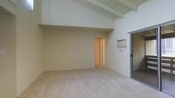 This single-floor space offers a clean, functional layout highlighted by a standout architectural feature: vaulted ceilings with exposed wooden beams painted white, lending both height and character to the main living area. The generous ceiling volume creates an open, airy atmosphere that makes the space feel expansive. Soft, neutral cream-toned walls complement the light-colored carpeted flooring throughout, providing a calm and versatile backdrop well-suited to a variety of furnishing styles. A wall-mounted air conditioning unit ensures climate control within the main living area. A sliding glass door on one side of the room provides direct access to the outdoors, allowing for natural light and easy indoor-outdoor flow. An interior doorway leads to an additional room or hallway, suggesting a practical, connected layout beyond the main living space. The raised ceiling section transitions to a slightly lower area near the rear of the room, adding architectural dimension. Overall, the space presents a straightforward yet charming interior defined by its notable beamed ceiling, neutral palette, and well-considered access points.