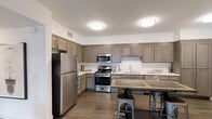 This single-floor space is thoughtfully designed to blend functionality with a warm, welcoming aesthetic. The kitchen serves as a central highlight, showcasing an expansive layout with abundant cabinetry finished in a rich taupe tone. Upper and lower cabinets provide generous storage throughout, complemented by clean white countertops that offer ample workspace for cooking and meal preparation. A full suite of stainless steel appliances includes a gas range with oven, an over-the-range microwave, a dishwasher, and a large refrigerator, equipping the space for everyday use. The deep single-basin sink is positioned beneath a dedicated cabinet section, adding to the kitchen's organized flow. Durable wood-look flooring runs throughout, lending warmth and continuity to the space. Flush-mount ceiling light fixtures provide consistent, bright illumination across the room. A kitchen island or bar-height table with industrial-style metal stools anchors the dining area, creating a casual spot for meals or conversation. The overall ambiance is modern yet comfortable, with neutral tones and practical finishes that make this a highly livable and versatile space.