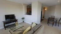 This single-floor residence is designed with an open-concept layout that seamlessly connects the living, dining, and kitchen areas. The living room is carpeted and furnished with a plush sofa and a glass-top coffee table, creating a comfortable and welcoming atmosphere. A wall-mounted TV setup anchors one side of the room, while a decorative potted plant adds a touch of greenery.

The kitchen is accessible via a distinctive pass-through window framed by a dark granite countertop, offering a convenient connection to the main living space. Warm wood cabinetry and stainless steel appliances, including a built-in microwave, are visible through the opening, giving the kitchen a functional yet stylish character.

Adjacent to the living room, the dining area accommodates a dark wood table and chairs, ideal for everyday meals or entertaining. Neutral beige walls and consistent carpeting throughout create a cohesive and understated aesthetic. A hallway fitted with a wall sconce leads further into the residence, hinting at additional private spaces beyond. The overall ambiance is warm, practical, and well-organized, making efficient use of the single-floor footprint.