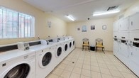 This shared laundry facility is a clean, functional space designed to efficiently serve a multi-unit community. The room is finished with light beige walls and durable ceramic tile flooring throughout, creating a bright and easy-to-maintain environment. Overhead fluorescent lighting ensures the space is well-lit at all times.

Along one wall, a row of commercial-grade front-load washing machines provides ample capacity for residents, while an adjacent wall houses a bank of stacked dryer units, offering an efficient use of vertical space and substantial drying capacity. A utility sink with under-counter cabinetry is available for hand-washing or pre-treating garments, and a fold-down laminate table provides a convenient surface for sorting and folding laundry.

A small seating area with two chairs and a waste bin offers a comfortable spot to wait during laundry cycles. Informational signage and laundry guidelines are posted on the walls for easy reference. A security camera is mounted on the ceiling, contributing to the overall safety of the space. Natural light filters in through a window fitted with blinds, complementing the overhead lighting. A fire extinguisher and clearly marked exit door ensure the facility meets safety standards. The space is practical, orderly, and thoughtfully arranged for daily communal use.