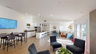 This single-floor space presents a thoughtfully designed open-plan layout that seamlessly connects the kitchen, dining, and multiple lounge areas into one cohesive and inviting environment. Light hardwood flooring runs throughout, reflecting generous natural light that streams in through large French doors and plantation-shuttered windows, creating a warm and airy atmosphere.

The kitchen is well-appointed with white shaker cabinetry, dark stone countertops, stainless steel appliances including a French door refrigerator, dishwasher, and built-in microwave, along with a central island with bar seating. Open shelving adds a contemporary touch and provides additional display and storage space.

The main lounge area features a striking stone-clad fireplace as a focal point, flanked by French doors that open to the outdoors. Comfortable seating arrangements — including upholstered sofas, mid-century accent chairs in deep blue tones, and a live-edge wood coffee table — define distinct relaxation zones within the open space.

A secondary lounge area includes a large wall-mounted display screen, a bar-height work or dining table, and additional cabinetry, offering a versatile space for gatherings or focused activities. A dining table with chairs provides a dedicated area for meals. Recessed lighting and vaulted ceilings in select areas further enhance the sense of openness and sophistication throughout.