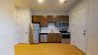 Located at 1341 Norton Ave in Columbus, Ohio's Grandview Heights area, this single-floor unit offers a clean, modern design throughout. The kitchen is a standout feature, equipped with a full suite of stainless steel appliances including a refrigerator, electric range, built-in microwave, and dishwasher. Rich walnut-toned cabinetry extends to the upper walls, complemented by a light quartz countertop and a subway tile backsplash that adds a contemporary touch. Light hardwood-style flooring runs consistently throughout the unit, creating a cohesive and airy feel. A large window in the kitchen area allows natural light to pour in, offering views of the surrounding neighborhood. The living area is open and versatile, with neutral light-gray walls and a warm accent of yellow near the ceiling, adding a subtle pop of color. A sliding glass-panel closet provides practical storage. The bathroom is neatly appointed with a wood-tone vanity, white countertop, and clean white walls, maintaining the unit's consistent modern aesthetic. The overall layout, as seen in the dollhouse view, is efficient and well-organized, with clearly defined zones for living, cooking, and bathing.