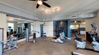 This fitness center is thoughtfully designed to support a comprehensive workout routine in a clean, modern environment. The space features a combination of cardio equipment, including stair climbers and other aerobic machines, alongside a multi-station strength training unit. A dedicated free weights area with a full dumbbell rack is positioned near floor-to-ceiling mirrors, allowing users to monitor their form during exercise. The flooring consists of durable, patterned carpet tiles that provide both comfort and traction throughout the space. Overhead, a ceiling fan ensures consistent air circulation, complemented by track lighting that delivers bright, even illumination across the room. Crown molding and crisp white trim add a refined architectural touch, while a deep blue accent wall creates a bold visual focal point that distinguishes the cardio zone. Ventilation is well-addressed with multiple ceiling vents, maintaining a comfortable climate during use. The mirrored walls enhance the sense of openness and allow for easy visual monitoring of the entire room. Overall, the fitness center combines practical functionality with polished design elements, offering an inviting and motivating environment for users of all fitness levels.