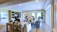 This single-story residence offers a refined and thoughtfully designed interior centered around a generous open-plan living space. The layout seamlessly connects multiple living zones, creating a sense of flow and openness throughout. Wide-plank hardwood flooring runs continuously across the space, anchoring the warm yet sophisticated aesthetic.

The main living area is highlighted by a classic fireplace with a decorative mantel, serving as a natural focal point. A built-in shelving and media unit lines one wall, offering both storage and display space, currently styled with curated décor pieces. Recessed lighting is fitted throughout the ceiling, complemented by elegant crown molding that frames each zone with architectural detail.

Comfortable seating arrangements, including plush sofas and accent chairs, are arranged over a patterned area rug to define the central gathering space. A wooden console table adorned with decorative sculptures, including a distinctive carved horse figurine, adds artistic character to the room.

Large windows along the far wall invite an abundance of natural light, creating a bright and airy ambiance. A dining area positioned near the windows takes advantage of the natural illumination, making it an inviting space for meals. The overall atmosphere is polished and welcoming, blending classic architectural details with contemporary furnishings.