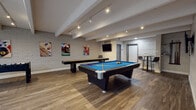 Located at 11100 E Dartmouth Ave in Denver, Colorado, this single-floor community game and recreation room offers a dynamic and social environment designed for entertainment and leisure. The space is defined by its exposed beam ceiling with track lighting and recessed fixtures that cast a warm, inviting glow throughout the room. Durable wood-look flooring runs the length of the space, complementing the painted white brick accent walls adorned with vibrant sports-themed artwork.

The main game area is generously sized to comfortably accommodate a full-size pool table with a vivid blue felt surface, a long shuffleboard table, and a foosball table — providing a variety of recreational options for guests. Multiple wall-mounted flat-screen televisions are positioned throughout the room for easy viewing from various angles, making it equally suited for watching sports or casual gatherings.

Adjacent to the game area is a well-equipped kitchenette featuring a white quartz peninsula with an undermount sink and bar seating, open counter space with a coffee station and beverage appliances, a beverage refrigerator, and a tiled backsplash. High-top bar tables and seating are also available, rounding out the space as a versatile hub for socializing and recreation.