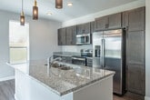 This well-designed single-floor home offers a cohesive, modern aesthetic carried throughout each room. The open-concept kitchen serves as a central highlight, featuring gray-toned shaker-style cabinetry that extends to the ceiling, granite countertops, and a full suite of stainless steel appliances including a refrigerator, range, and microwave. A spacious island with a built-in sink and elegant pendant lighting anchors the kitchen, making it as functional as it is visually appealing. Hardwood-style flooring flows consistently across the living areas, bedrooms, and hallways, tying the home together with a warm, uniform look. The living area is generously sized, bathed in natural light through multiple windows and a sliding glass door fitted with vertical blinds that open to outdoor views. A ceiling fan adds comfort to the space. The bedrooms are bright and neutral, each featuring large windows with blinds that invite natural light. The bathrooms are finished with granite-topped dark wood vanities, framed mirrors, modern light fixtures, and matte black hardware accents. One bathroom includes a soaking tub, while both maintain a clean, contemporary feel. Throughout, the home is finished with crisp white trim, neutral gray walls, and recessed lighting, creating a bright and inviting atmosphere.