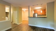 This single-floor space showcases a thoughtfully designed open-concept layout that seamlessly connects the kitchen and living areas. The kitchen is a standout feature, equipped with warm wood cabinetry, dark countertops, and stainless steel appliances, including a full-size refrigerator. A raised breakfast bar serves as a natural divider between the kitchen and the adjoining living space, making it ideal for both casual dining and entertaining. Elegant pendant lighting hangs above the bar, complementing the track lighting overhead and adding a modern touch to the room's ambiance. The living area flows naturally from the kitchen, offering an open and airy feel enhanced by soft, neutral wall tones in warm green and white hues. Rich hardwood flooring runs throughout the space, providing a cohesive and inviting aesthetic. A structural column adds an architectural element to the interior, while a white paneled door leads to an additional room or entryway, hinting at further private spaces beyond the main living area. Recessed and flush-mount ceiling lighting ensures the entire space is well-lit, creating a bright and welcoming environment throughout.