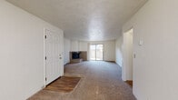 This single-floor residence offers a functional and straightforward layout across several well-defined living spaces. The expansive living room serves as the heart of the home, highlighted by a corner brick fireplace with a raised hearth that adds a classic, cozy focal point. Plush wall-to-wall carpeting runs throughout the living areas and bedrooms, while a sliding glass door floods the space with natural light and provides direct access to an outdoor balcony or patio enclosed by a railing. The kitchen is equipped with wood-toned cabinetry, yellow laminate countertops, a white electric range with an overhead ventilation hood, and a window that allows for additional natural light. A carpeted hallway connects the main living area to the private quarters, where bedrooms feature textured ceilings, neutral walls, and ample closet space with sliding panel doors. The full bathroom is finished with wood-look vinyl flooring, a single-sink vanity with wood cabinetry, a toilet, and a tub-shower combination with a white surround. Throughout the residence, textured ceilings, white walls, and consistent neutral tones create a clean, understated aesthetic.