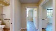 This single-floor unit is designed with a clean, functional layout that makes efficient use of its space. Throughout the unit, light-toned luxury vinyl plank flooring runs continuously, providing a cohesive and low-maintenance foundation that ties each area together. Walls are painted in a warm, muted yellow-beige tone, lending a neutral and welcoming ambiance to every room.

A central hallway connects the unit's various spaces, offering clear sightlines and easy circulation. The kitchen area is equipped with white cabinetry — including both upper and lower storage units — a compact sink, a refrigerator, and space for a microwave, offering practical functionality within a modest footprint. A tall storage cabinet provides additional organizational capacity.

The bathroom features a walk-in shower with a handheld showerhead and chrome fixtures, complemented by the same consistent flooring and neutral wall tones found throughout the unit.

One of the standout spaces is a corner room benefiting from dual-aspect windows that frame lush, green tree canopy views, allowing abundant natural light to fill the room. A baseboard heater ensures comfort year-round. Additional rooms provide quiet, versatile spaces suited for various uses. Overall, the unit presents a tidy, move-in-ready environment with a calm and understated aesthetic.