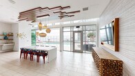 Located at 707 E Ocean Blvd in Long Beach, California, Altitude 155 is an elevated amenity space that seamlessly blends modern interior design with exceptional outdoor living. The spacious living area is defined by soaring ceilings adorned with a striking geometric wood slat installation, complemented by sculptural pendant lighting in warm amber tones. Large floor-to-ceiling glass walls and doors flood the interior with natural light while framing direct views of the outdoor pool and surrounding skyline. Light-toned herringbone-patterned tile flooring and a long white communal table anchor the central gathering space, while a rustic stacked-wood bar counter adds an organic, textural contrast. A separate lounge area features built-in gray banquette seating with gold accent pillows and warm wall sconces, creating an intimate retreat within the broader space. Outside, multiple terraces wrap around the building, offering residents a pool deck with lounge chairs, a built-in BBQ station clad in blue tile, a plunge pool bordered by river rock and tropical plantings, and al fresco dining areas. Glass railings ensure unobstructed, panoramic views of the Long Beach harbor, marina, and downtown skyline, making the outdoor spaces as compelling as the interior.