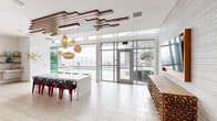 Located at 707 E Ocean Blvd in Long Beach, California, Altitude 155 is an elevated amenity space that seamlessly blends modern interior design with exceptional outdoor living. The spacious living area is defined by soaring ceilings adorned with a striking geometric wood slat installation, complemented by sculptural pendant lighting in warm amber tones. Large floor-to-ceiling glass walls and doors flood the interior with natural light while framing direct views of the outdoor pool and surrounding skyline. Light-toned herringbone-patterned tile flooring and a long white communal table anchor the central gathering space, while a rustic stacked-wood bar counter adds an organic, textural contrast. A separate lounge area features built-in gray banquette seating with gold accent pillows and warm wall sconces, creating an intimate retreat within the broader space. Outside, multiple terraces wrap around the building, offering residents a pool deck with lounge chairs, a built-in BBQ station clad in blue tile, a plunge pool bordered by river rock and tropical plantings, and al fresco dining areas. Glass railings ensure unobstructed, panoramic views of the Long Beach harbor, marina, and downtown skyline, making the outdoor spaces as compelling as the interior.
