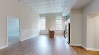 This single-floor unit blends historic architectural character with modern finishes throughout its well-organized layout. The main living and kitchen area immediately draws attention with its striking pressed-tin ceiling, a decorative coffered design that adds depth and elegance to the space. Tall, arched windows flood the room with natural light and offer views of the surrounding brick exteriors, reinforcing the building's classic urban character. Wainscoting panels line the lower walls, complementing the warm hardwood-style flooring that runs throughout the main area.

The kitchen is thoughtfully designed with white shaker cabinetry, light countertops, and a full suite of stainless steel appliances, including a range, dishwasher, microwave, and refrigerator. A central island provides additional prep and seating space, with a peninsula-style counter further separating the kitchen from the living area.

The bedrooms feature neutral carpet, ample wall space, and windows that allow natural light to filter in. Each room connects conveniently to the rest of the unit through well-placed doorways visible in the open floor plan.

Two full bathrooms are finished with classic white subway tile surrounding the tub-shower combinations, neutral floor tile, and clean white vanities, offering a crisp, timeless aesthetic throughout.