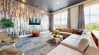 This contemporary amenity lounge spans a generous single floor, offering a seamlessly connected dining and living area complemented by a hallway and an expansive outdoor terrace. The combined dining and living space is defined by rich design details, including a dramatic dark ceiling with recessed lighting and a bold, abstract-patterned accent wall in the lounge area, where a large sectional sofa and plush leather armchairs create inviting conversation zones. Floor-to-ceiling windows flood the interior with natural light while framing sweeping outdoor views of the surrounding cityscape and greenery. A dedicated dining zone features a substantial wood communal table beneath striking oversized pendant lights with gold-interior shades, paired with a sleek wet bar finished in chevron-patterned metallic tile and stone countertops. Throughout, light wood-plank flooring and patterned carpet tile provide textural contrast. The hallway connects the interior to the outdoor terrace, which is finished with large-format light tile flooring under a warm yellow ceiling with recessed lighting. The terrace accommodates bar-height seating and lounge furniture, offering open-air views of the treetops and city skyline beyond a black steel railing. The overall design combines modern sophistication with a warm, welcoming ambiance.