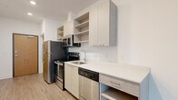 This single-floor unit showcases a clean, contemporary design characterized by crisp white walls, recessed lighting, and wood-look plank flooring that runs consistently throughout the space, creating a cohesive and modern aesthetic.

The kitchen is thoughtfully designed along a single wall, featuring light wood cabinetry paired with open shelving for practical storage and visual appeal. Stainless steel appliances include a refrigerator, electric range, built-in microwave, and dishwasher. Light-toned quartz countertops extend generously along the workspace, complementing the cabinetry's natural finish.

The layout is organized along a central corridor that connects the kitchen and living areas to the private rooms. The main living area benefits from a large window that draws in natural light and offers views of surrounding greenery, creating an open and airy atmosphere. The room's clean lines and neutral palette allow for versatile furnishing arrangements.

Private rooms are well-proportioned with white walls, wood-look flooring, and individual doors. The bathroom features a walk-in glass-enclosed shower with white subway tile walls and a pebble-tile floor, paired with a light wood vanity, white countertop, and a large mirror, maintaining the unit's consistent modern design language throughout.