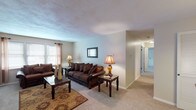Located in Camillus, NY, this single-floor residence offers a well-organized layout designed for comfortable everyday living. The living room serves as the central gathering space, featuring wall-to-wall carpeting, neutral-toned walls, and multiple windows that allow natural light to filter in through sheer curtains and blinds. The open flow connects seamlessly to a dedicated dining area, which is anchored by a pendant chandelier overhead and provides ample space for a full dining set — ideal for both casual meals and entertaining.

The kitchen is equipped with white cabinetry, dark granite-style countertops, a stainless steel range, and hardwood-style flooring, offering a practical and clean cooking environment. A hallway connects the living spaces to the private areas of the home, including a carpeted bedroom with a closet, wall-mounted air conditioning unit, and windows with blinds for light control. A second room with cool gray-toned walls and carpeting offers flexibility as an additional bedroom or office. The full bathroom features a classic white tiled tub and shower combination. Throughout the residence, a consistent neutral color palette and carpet flooring create a cohesive and inviting atmosphere.