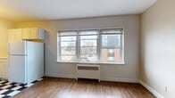 Located on N Prospect Ave in Milwaukee's vibrant East Side neighborhood, this single-floor unit offers a practical and functional layout with several notable features throughout. Wood-style flooring flows consistently across the main living areas, providing a warm and cohesive aesthetic.

The living area is generously proportioned, with neutral-toned walls and a bank of large windows fitted with roller blinds. A radiator beneath the windows provides heating, while the elevated position of the unit affords views of the surrounding Milwaukee cityscape, including nearby mid-rise buildings and street activity below.

Adjacent to the living space, the kitchen is distinguishable by its classic black-and-white checkered tile flooring, yellow-painted upper walls, and white cabinetry. A full-size refrigerator and additional appliances are included. The kitchen also benefits from window access, maintaining a bright atmosphere.

A dedicated closet space off the main living area offers additional storage. The bathroom features a tub and shower combination with a striped curtain, classic mosaic-patterned floor tile in neutral tones, and clean white walls, reflecting a straightforward, functional design. Overall, the space offers a bright, urban feel suited to everyday living.