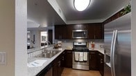 The Ashford A5 is a thoughtfully designed 2-bedroom, 2-bathroom residence situated on a single floor, offering a cohesive and functional living environment. The kitchen serves as a standout feature of the home, showcasing rich dark espresso cabinetry with sleek hardware that provides ample storage space. Granite-style countertops extend generously across the workspace, complemented by a classic mosaic tile backsplash that adds visual texture and depth. Stainless steel appliances, including a gas range, built-in microwave, and dishwasher, give the kitchen a contemporary, polished look. A double stainless steel sink is positioned along the counter with easy access to the adjacent workspace. The kitchen opens toward a connected living area, allowing for a natural flow between spaces and making the layout ideal for everyday living and entertaining. Light wood-tone flooring runs throughout, providing warmth and continuity across the open floor plan. Flush-mount ceiling lighting provides consistent illumination throughout the kitchen. The overall design balances modern aesthetics with practical functionality, creating a comfortable and stylish living environment in The Ashford A5.