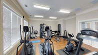 This fitness center offers a comprehensive workout environment designed to accommodate a range of exercise needs. The space features hardwood-style flooring throughout, providing a clean and durable surface ideal for both equipment use and open floor exercises, complemented by a yoga mat for stretching or low-impact workouts. Bright fluorescent lighting spans the ceiling, ensuring the room is well-lit and energizing at any time of day. Natural light also filters in through window blinds along one wall, adding a welcoming atmosphere to the space. The room is outfitted with an array of cardio equipment, including elliptical trainers, a stationary upright bike, and additional cycling-style machines. Strength training is well represented with multiple cable and weight machines, a bench press setup, and a dumbbell rack stocked with a range of free weights. Crown molding along the ceiling adds a subtle architectural detail, lending a polished finish to the room. The layout is thoughtfully organized to allow users to move freely between stations, making it a functional and accessible facility for a variety of fitness routines.