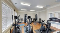 This fitness center offers a comprehensive workout environment designed to accommodate a range of exercise needs. The space features hardwood-style flooring throughout, providing a clean and durable surface ideal for both equipment use and open floor exercises, complemented by a yoga mat for stretching or low-impact workouts. Bright fluorescent lighting spans the ceiling, ensuring the room is well-lit and energizing at any time of day. Natural light also filters in through window blinds along one wall, adding a welcoming atmosphere to the space. The room is outfitted with an array of cardio equipment, including elliptical trainers, a stationary upright bike, and additional cycling-style machines. Strength training is well represented with multiple cable and weight machines, a bench press setup, and a dumbbell rack stocked with a range of free weights. Crown molding along the ceiling adds a subtle architectural detail, lending a polished finish to the room. The layout is thoughtfully organized to allow users to move freely between stations, making it a functional and accessible facility for a variety of fitness routines.