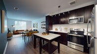 This single-floor residence showcases a thoughtfully designed open-concept layout that seamlessly connects the kitchen, dining, and living areas. The kitchen is a standout feature, outfitted with rich dark espresso cabinetry, white quartz countertops, a white subway tile backsplash, and a full suite of stainless steel appliances including a refrigerator, range, microwave, and dishwasher. A freestanding kitchen island provides additional prep and seating space, while track lighting overhead adds a modern touch. The dining area flows naturally into the living room, which is bright and inviting thanks to large windows that allow generous natural light and views of the surrounding greenery. The living room features warm wood-look flooring, contemporary furnishings, and a dedicated media area. The bedroom offers a serene retreat, highlighted by a bold navy accent wall, a large window with outdoor views, soft carpeting, and ample room for furnishings. A closeted area provides convenient storage access to the main living spaces. The bathroom is finished with neutral tile surround, a soaking tub and shower combination, a dark-cabinet vanity with white quartz countertop, and a large mirror. Throughout the residence, a consistent palette of warm neutrals, dark wood tones, and crisp white surfaces creates a cohesive and modern aesthetic.