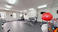 Located on Brooklyn Ave in Evansville, Indiana, this single-floor fitness center offers a comprehensive workout environment designed to accommodate a wide range of exercise needs. The space is organized into distinct functional zones, making it easy to transition between cardio, strength training, and flexibility work.

The cardio area is outfitted with a treadmill, elliptical trainer, and stationary bike, all positioned to allow comfortable spacing between users. A wall-mounted flat-screen television is positioned nearby, adding convenience during longer sessions. The strength training section features multiple selectorized cable and resistance machines, providing options for targeting various muscle groups. A dedicated free weight area includes a multi-tier dumbbell rack stocked with a full range of weights, paired with an adjustable weight bench. Stability balls of varying sizes round out the functional fitness offerings.

Full-length wall mirrors span one side of the room, visually expanding the space and allowing users to monitor their form. Durable rubber flooring covers the entire area, providing both safety and shock absorption. Bright recessed fluorescent lighting ensures even illumination throughout, while several windows along the walls allow natural light to supplement the interior fixtures. The walls are finished in a clean, neutral tone, contributing to a bright and motivating atmosphere.