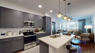 This single-floor residence showcases a thoughtfully designed open-concept layout with cohesive modern finishes throughout. The kitchen serves as the centerpiece of the living area, featuring floor-to-ceiling gray shaker cabinetry, white quartz countertops, and a full suite of stainless steel appliances including a range, microwave, and refrigerator. A substantial kitchen island with a striking blue base provides additional prep space and seating, illuminated by a trio of globe pendant lights in warm brass tones. Rich hardwood flooring flows throughout the main living areas, grounding the open-concept design.

The living room receives generous natural light through large windows dressed with floor-length drapes, offering outdoor views of surrounding greenery. The space accommodates comfortable seating and features hardwood floors that enhance the room's warmth.

The bedrooms are carpeted for comfort and each include ceiling fans, natural light from large windows with blinds, and ample room for furnishings. The bathroom features a wide vanity with dark navy cabinetry, a backlit mirror, quartz countertops, wood-look tile flooring, and a full tub-shower combination. Recessed lighting throughout the residence ensures consistent, well-distributed illumination across all spaces.