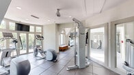 This well-appointed fitness center in Dunwoody, Georgia offers a comprehensive workout environment spread across a single floor. The facility is divided into two dedicated exercise rooms, a living/lounge area, and an entrance, creating a logical flow for users moving through the space.

The primary exercise room is bright and airy, featuring large curved windows that flood the area with natural light and offer views of the surrounding tree-lined streetscape. This room houses cardio equipment including treadmills and an elliptical trainer, each positioned to take advantage of the outdoor views. Wall-mounted flat-screen TVs provide entertainment during workouts, and full-length mirrors expand the sense of space. Durable rubber-tile flooring covers the exercise areas, while large-format tile defines the transitional zones.

The secondary exercise room offers a more intimate setting with bold blue accent walls and recessed lighting. It is equipped with strength-training machines, a cable weight system, a stability ball, free weights, and a workout bench — catering to a full range of resistance training needs.

A connecting corridor with additional strength equipment links the two rooms, while a cozy lounge/living area with a wooden bench provides a space to rest. The entrance is clean and functional, with glass-panel doors offering a welcoming transition into the facility. Throughout, the design balances practicality with an inviting, modern aesthetic.
