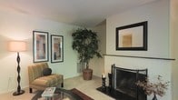 This single-floor space presents a comfortable and well-appointed living area that balances functionality with classic décor. The living room serves as the focal point, anchored by a brick fireplace fitted with a black metal screen, adding a traditional and inviting element to the space. A large, lush indoor tree in a woven basket planter stands prominently nearby, bringing a natural, organic touch to the interior. The walls are painted in a soft, neutral tone that contributes to the room's calm and welcoming atmosphere. A framed mirror is mounted above the fireplace surround, enhancing the sense of depth in the room. Two framed artworks are displayed side by side on an adjacent wall, adding a curated aesthetic to the space. Seating includes a striped upholstered armchair with a coordinating throw pillow, positioned alongside a glass-topped coffee table. Light-toned tile flooring runs throughout, accented by a patterned area rug that defines the seating zone. Decorative candle holders are arranged near the fireplace, completing the warm and tasteful presentation of this single-level home.