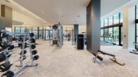 Located at 45 Northeast 27th Street in Miami's vibrant Edgewater district, this professionally outfitted fitness center offers an expansive, single-floor layout designed to accommodate a full range of workout disciplines. The gym's modern architecture is defined by soaring ceilings with recessed lighting, large floor-to-ceiling windows that flood the space with natural light and provide views of lush outdoor greenery, and sleek dark accent columns that frame the open floor plan.

The facility is comprehensively equipped with a premium lineup of Life Fitness machines, including treadmills, stair climbers, ellipticals, and stationary cycling bikes. A dedicated strength training area features a multi-station cable machine, adjustable benches, and a well-stocked dumbbell rack ranging from light to heavy weights. Functional training zones provide ample open floor space for stretching and bodyweight exercises.

Full-length mirrors line the walls throughout, visually expanding the already generous footprint and allowing users to monitor their form. The neutral-toned, cushioned flooring adds comfort underfoot while complementing the clean, contemporary aesthetic of warm whites and charcoal grey accents. A built-in water fountain station ensures convenient hydration. The overall ambiance is bright, airy, and professional, creating an inviting environment for focused fitness pursuits.