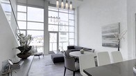 This two-story space makes an immediate impression with its dramatic double-height ceilings and expansive floor-to-ceiling glass walls that flood the interior with natural light. The oversized windows frame panoramic views of the surrounding cityscape and hillside, creating a seamless connection between the interior and the urban environment beyond.

The main living area is designed with a clean, contemporary aesthetic, anchored by a sophisticated pendant light fixture featuring multiple cylindrical glass shades suspended at varying heights — a striking focal point that complements the vertical scale of the room. The open layout accommodates both a relaxed lounge area and a dedicated dining space, with modern furnishings in a neutral palette of whites, grays, and charcoals that reinforce the space's minimalist sensibility.

Wide-plank hardwood flooring runs throughout, adding warmth and texture to the otherwise crisp, white interior. The white walls serve as a refined backdrop for curated artwork. A private balcony accessible through the glass facade extends the living space outdoors, enhancing the sense of openness. The two-floor layout offers versatility and an elevated sense of scale rarely found in urban settings.