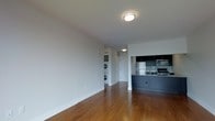 This single-floor residence offers a thoughtfully designed open-concept layout that seamlessly integrates the living area and kitchen. Rich hardwood flooring flows throughout the space, lending warmth and continuity to the interior.

The kitchen is a standout feature, equipped with stainless steel appliances including a refrigerator, microwave, and range. Dark cabinetry provides a modern contrast against a classic tile backsplash, while the kitchen island with its clean countertop offers both additional prep space and a natural divider between the cooking and living areas.

The open living area is bright and spacious, with clean white walls and flush ceiling lighting that create a neutral, welcoming ambiance. The layout allows for flexible furniture arrangement to suit a variety of living styles.

A particularly practical feature of this residence is the in-unit stacked washer and dryer, neatly integrated into a dedicated alcove, providing added convenience without occupying excessive space. A doorway at the rear of the living area leads to additional rooms beyond, suggesting well-organized private quarters. Overall, the space strikes a balance between contemporary aesthetics and functional everyday living.