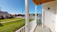 This single-story community space is thoughtfully designed to offer a welcoming and refined atmosphere for gathering and relaxation. The interior is characterized by soaring ceilings, crisp white walls, and dark-trimmed French doors that frame views between rooms and allow abundant natural light to flow throughout.

The main lounge area serves as the heart of the space, furnished with plush cream sofas, wingback chairs, and soft accent pillows in aqua and gold tones. Elegant multi-bulb chandeliers hang overhead, adding a warm, sophisticated glow. A striking floor-to-ceiling stone fireplace with a rustic wood mantel anchors one end of the room, providing both a visual focal point and a cozy gathering spot.

French doors open directly to an outdoor covered porch, which features white-painted columns and railings beneath a warm wood-paneled ceiling. The porch overlooks well-maintained green lawns, a parking area, and a residential neighborhood backdrop under open skies — ideal for enjoying fresh air and natural surroundings.

An adjoining area provides a more intimate setting with a dining table and additional seating, connected seamlessly to the main lounge through open archways. The grand entry features tall dark wood doors with transom windows, creating a formal and inviting arrival experience. Throughout, the neutral color palette, carpeted floors, and carefully curated furnishings contribute to a cohesive, upscale ambiance.