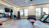 This fitness center is thoughtfully designed to support a comprehensive workout experience. The space features a distinctive hexagonal-patterned flooring that adds a modern aesthetic throughout. Recessed lighting and crown molding detail the ceiling, lending the room a polished, upscale feel. Multiple windows along the exterior wall allow natural light to fill the space and provide views of the surrounding outdoor environment.

The cardio zone is well-stocked with several elliptical trainers, stationary bikes, and treadmills, each equipped with individual display screens. A dedicated strength training area houses a cable machine and a leg press/extension unit with a red padded seat, complemented by a flat utility bench. A two-tier dumbbell rack lines one wall, offering a wide range of free weight options.

Large floor-to-ceiling mirrors span one wall, visually expanding the space and allowing users to monitor their form during exercise. Wall-mounted flat-screen televisions are positioned throughout for entertainment. Near the entrance, a built-in storage cabinet with cubbies provides convenient organization. A fitness center information board is also posted near the entry. The overall ambiance is clean, bright, and welcoming, making it a functional and inviting environment for exercise.