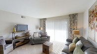 This 2-bedroom, 1-bathroom single-floor residence offers a well-designed layout suited for comfortable everyday living. The living room serves as the centerpiece of the home, featuring plush carpeted flooring and a neutral color palette that creates a calm, inviting atmosphere. A large sliding glass door flanked by decorative patterned curtains floods the space with abundant natural light, enhancing the open, airy feel of the room. The living area comfortably accommodates a full seating arrangement, including a loveseat and additional seating, complemented by a rustic wooden storage trunk that doubles as a coffee table. A media console along the adjacent wall provides space for a flat-screen television and decorative accents. Ceiling-mounted ventilation ensures year-round comfort throughout the space. The clean white ceilings and light-toned walls contribute to a bright, cohesive interior aesthetic. With its practical layout and tasteful design details, this single-story residence offers a functional and comfortable living environment suitable for a range of lifestyles.