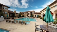 This outdoor pool area offers a well-appointed, resort-inspired retreat designed for relaxation and social gathering. The centerpiece is a generously sized swimming pool with clear blue water, complemented by a separate hot tub/spa positioned along the pool deck. The surrounding concrete deck provides ample space for rows of lounge chairs and patio seating, making it ideal for sunbathing or casual gatherings.

A covered pergola structure supported by substantial stone columns serves as a shaded lounge area, furnished with cushioned outdoor sofas and accent tables. String lights are strung across the pergola's wooden beams, adding ambiance during evening hours. An outdoor fireplace with decorative ironwork detailing is situated beneath the pergola, offering warmth and a focal point for cooler days.

Additional open-air seating areas with tables, chairs, and teal market umbrellas are scattered throughout the courtyard, providing flexible gathering spots. The space is enclosed by iron fencing with rock landscaping, evergreen trees, and potted plants that add natural texture and color. The surrounding multi-story buildings frame the courtyard on multiple sides, creating an intimate, community-centered atmosphere with open sky views above.
