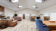 Located at 2895 E Charleston Blvd in Las Vegas, NV, this single-floor commercial space offers a generously sized, open-plan layout designed for both comfort and functionality. The standout architectural feature is the dramatic vaulted ceiling, which rises impressively overhead and is punctuated by multiple skylights that flood the interior with abundant natural light throughout the day. Ceiling fans are strategically positioned to complement the airy atmosphere.

The main area serves as a welcoming lounge and gathering space, furnished with plush brown leather sectional sofas accented by vibrant blue and red throw pillows, arranged atop colorful area rugs over light wood-tone luxury vinyl plank flooring that runs throughout the entire space. A mounted flat-screen television, rustic open shelving units, and decorative lanterns contribute to the warm, eclectic aesthetic. An exposed brick accent feature adds character to one wall, while an inspirational wall decal reading "Some people dream of success, others make it happen" reinforces the space's motivational atmosphere.

A sliding barn door separates a secondary area that includes a dining table with seating and a dedicated workstation zone with a conference-style desk. The space also includes a fully equipped kitchen featuring white shaker cabinetry, stainless steel appliances, a gas range, and a mosaic tile backsplash. French doors and horizontal blinds provide privacy while maintaining a bright, open feel throughout.