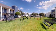 Located at 2301 Windchester St in Pearland, Texas, this single-story unit offers a thoughtfully designed interior complemented by well-maintained community outdoor amenities. The living area immediately draws attention with its soaring vaulted ceilings, which lend an open, airy quality to the space. A tile-surround fireplace with a white mantel serves as a natural focal point, adding warmth and character to the room. Dark hardwood-style flooring runs throughout, providing a rich contrast against the neutral, light-toned walls.

Large windows allow generous natural light to fill the living space, offering views of the surrounding greenery and neighborhood streetscape. The tall windows extend toward the upper portion of the vaulted ceiling in select areas, further enhancing the sense of vertical scale. A staircase with white railings and elegant arched upper windows adds architectural interest, while an in-unit laundry area is conveniently tucked beneath the stairwell.

Outdoors, the property features a well-kept grassy common area enclosed by chain-link fencing, complete with a dedicated dog park equipped with charming pet-themed metal sculptures, benches, and a fire hydrant — ideal for pet owners. The community setting is surrounded by mature trees beneath open Texas skies.