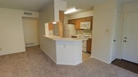 This single-floor unit is designed with an open-concept layout that seamlessly connects the kitchen and living areas, creating a cohesive and functional living space. The kitchen is well-appointed with warm medium-toned wood cabinetry offering generous storage, white appliances including a refrigerator, electric range, and over-the-range microwave, and light-colored countertops that brighten the space. A peninsula-style breakfast bar extends into the living area, complete with a sink, providing a convenient spot for casual dining or meal preparation while remaining connected to the rest of the space. The living area is finished with plush wall-to-wall carpeting in a neutral tone that adds warmth and comfort underfoot. The textured ceiling and soft, neutral wall colors throughout contribute to a calm, welcoming ambiance. A mirrored closet is visible along one wall, enhancing the sense of space and offering practical storage. The layout flows efficiently, making the most of the available footprint while maintaining an airy, open feel throughout the home.