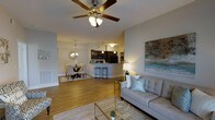 This single-floor residence offers a thoughtfully designed open-concept layout that connects the living, dining, and kitchen areas into one cohesive and functional space. The living room serves as the central gathering area, featuring warm hardwood-style flooring, a ceiling fan with integrated lighting, and ample room for comfortable furnishings. A large abstract wall canvas adds a decorative focal point, enhancing the room's contemporary aesthetic. The adjoining dining area is defined by an elegant chandelier and offers an intimate setting for meals. The kitchen is positioned behind a raised breakfast bar with seating for multiple guests, creating a natural divide while maintaining the open feel of the space. Dark cabinetry and black appliances give the kitchen a bold, modern contrast against the neutral-toned walls. Throughout the space, the neutral color palette of soft grays and warm whites creates a clean, inviting ambiance that feels both modern and livable. The consistent hardwood-style flooring flows seamlessly across all areas, tying the space together visually. Recessed lighting and pendant fixtures provide layered illumination throughout, complementing the overall warm and welcoming atmosphere of this well-designed residence.