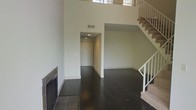 This two-story residence makes a striking first impression with its soaring double-height ceilings in the main living area, creating an expansive, airy atmosphere that feels open and grand. The main level showcases rich, dark hardwood flooring that flows seamlessly throughout the space, offering a bold contrast against the clean, neutral-toned walls.

The living room serves as the centerpiece of the main floor, anchored by a floor-to-ceiling tiled fireplace surround that adds texture and warmth to the space. A hallway connects the living area to additional rooms, recessed lighting guiding the way and ensuring a well-lit, functional layout. The open-concept design encourages natural movement between spaces.

A carpeted staircase with white balusters and a clean railing system rises from the main level to the second floor, where an upper landing overlooks the living area below, enhancing the sense of vertical scale. Windows positioned at the upper level allow natural light to filter in from above, brightening the interior throughout the day.

The overall design aesthetic is modern and understated, with a neutral palette, clean lines, and quality finishes throughout. The combination of dark flooring, white walls, and architectural volume gives this space a refined, move-in-ready appeal suited for a variety of furnishing styles.
