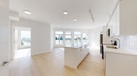 This thoughtfully designed 3-bedroom, 2-bathroom residence offers a clean, contemporary aesthetic across a single floor. The open-concept living and kitchen area forms the heart of the home, anchored by a generous central island with a smooth white countertop that doubles as a casual dining and prep space. The kitchen is well-appointed with high-gloss white cabinetry extending to the ceiling, a white subway tile backsplash, and a full suite of stainless steel appliances including a refrigerator, range, microwave, and dishwasher. Light hardwood flooring flows seamlessly throughout the entire unit, enhancing the bright, airy atmosphere. Large windows along the living area face outward to open sky views, flooding the space with natural light. Each of the three bedrooms is finished in crisp white with light hardwood floors and large windows that frame outdoor views of the surrounding cityscape. The two bathrooms mirror the home's minimalist style, featuring white floating vanities with undermount sinks, large mirrors, chrome fixtures, and combined tub-shower units. A clean hallway connects the private sleeping quarters, completing the efficient and well-organized floor plan.