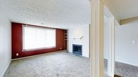 This single-floor space offers a comfortable and functional layout centered around a welcoming living area. The room is finished with wall-to-wall carpet that adds warmth underfoot and contributes to an overall cozy atmosphere. A standout feature is the classic fireplace framed by a clean white surround and mantel, serving as a natural focal point for the space. Flanking one side of the fireplace wall are built-in open shelves, offering both practical storage and display options. A bold deep-red accent wall adds a dramatic contrast to the otherwise neutral color palette, lending the room a distinct sense of character. Large windows dressed with vertical blinds span one wall, allowing generous natural light to fill the space and creating a bright, airy feel. The textured ceiling adds a traditional touch consistent throughout the room. A decorative column near the entryway subtly defines the transition into the living area, adding an architectural detail that enhances the overall structure of the space. Together, these elements create a living environment that balances classic design with everyday practicality.