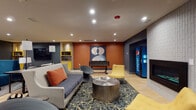 This expansive single-floor amenity lounge is thoughtfully designed to accommodate a variety of social and recreational activities. The space is divided into several distinct zones that flow seamlessly into one another across light wood-look tile flooring throughout.

The main lounge area features multiple seating arrangements, including a bold teal sofa, plaid upholstered chairs with yellow accent cushions, and a navy blue sectional grouping, all anchored by area rugs and coffee tables. A large-format wall-mounted television serves as the focal point of a dedicated media zone, ideal for group viewing.

A white textured tile fireplace wall creates a cozy retreat with neutral sofas and a paired coffee table arrangement nearby. Adjacent to this, a games corner offers a foosball table, dartboard, and an oversized Scrabble board, accompanied by a snack vending machine and beverage dispensers for convenience.

The full kitchen is well-appointed with stainless steel appliances—including a French door refrigerator, range, microwave, and dishwasher—complemented by white upper cabinetry, gray lower cabinets, a hexagonal tile backsplash, and a dark island with counter seating.

Bold orange accent walls, curated artwork, built-in shelving with yellow accents, and recessed lighting throughout contribute to a lively yet cohesive contemporary aesthetic.