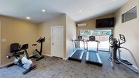 This residential community amenity space combines outdoor leisure and indoor fitness in a thoughtfully designed setting. The exterior showcases a Spanish-influenced architectural style, characterized by tiled rooflines, warm stucco facades, and wrought-iron staircases, all framed by vibrant tropical landscaping including towering palm trees and flowering shrubs that create an inviting curb appeal.

At the heart of the outdoor area lies a generously sized resort-style swimming pool with crystal-clear turquoise water, surrounded by a spacious concrete deck. Lounge chairs and a shaded canopy provide comfortable spots for relaxation, while secure fencing ensures safety around the pool perimeter.

The onsite fitness center is well-appointed and designed for a full-body workout experience. Large windows overlook the pool area, filling the space with natural light and outdoor views. The cardio section features multiple treadmills, an elliptical trainer, and a stationary bike. A dedicated strength training area is equipped with a professional-grade cable machine, a full rack of dumbbells, medicine balls, a weight bench, and wall-to-wall mirrors that amplify the sense of space. Wall-mounted televisions and recessed lighting complete the gym, making it a functional and motivating environment for residents.